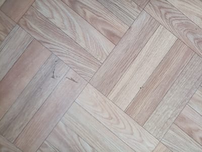 The image shows a close-up, overhead view of a light brown, wood-patterned floor with a herringbone design. The floor appears to be made of laminate or vinyl planks, with visible grain patterns and some darker lines indicating the seams between the planks. There are a few small, dark specks on the surface, possibly dust or minor imperfections. The lighting is even, casting no harsh shadows. The scene is indoors and sterile, with no context of the surrounding environment. There are no people or any other objects visible. The location is not specified in the image itself.