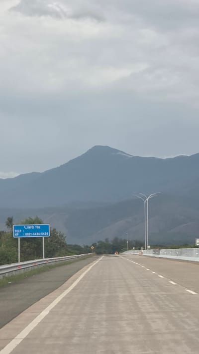 The image shows a highway adjacent to a mountain range under an overcast sky. The highway is grey, and a white line marks the edge. A blue sign with white text provides toll information, specifically a telephone number: 0821-6434-6434. The location is Baksukon, Indonesia. The environment appears humid, and no people are visible in the frame.