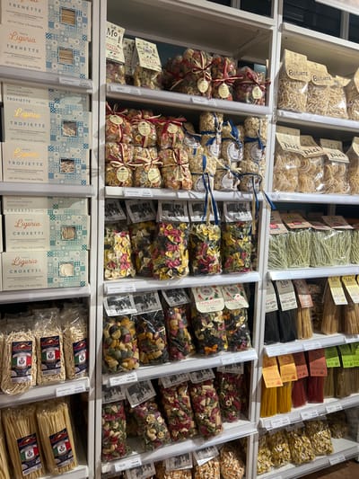 A well-organized grocery shelf displays various Italian pasta products. The top row features packaged pasta shapes like busiate, pici, and long strands. The second row contains creatively packaged pasta with ribbons and tags, alongside bags of colorful farfalle and other shapes. The bottom row presents neatly boxed "Liguria" pasta including trenette, trofiette, and croxetti, as well as additional bags of pasta. Brands like "Mo Morelli Italia" and "Mon" are visible, suggesting a focus on high-quality Italian goods.