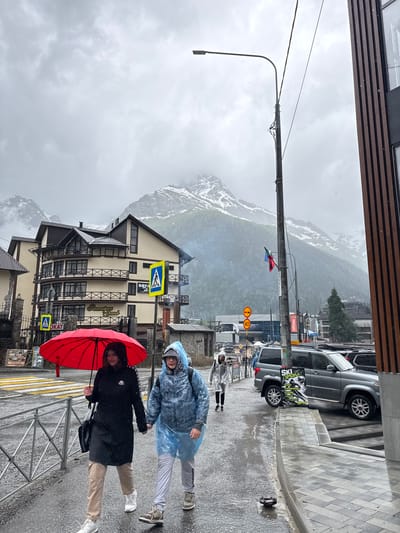 In Dombay, Russia, under a cloudy and rainy sky, two individuals are walking along a wet sidewalk. The person on the left holds a bright red umbrella, shielding themselves from the rain, while the person on the right wears a blue disposable poncho. They are walking hand-in-hand. Behind them, a multi-story hotel with balconies and dark windows stands prominently, adorned with the text "Ресторан Grand". Several road signs, including pedestrian crossing signs and a speed limit sign indicating 40, are visible. In the background, majestic, snow-capped mountains loom under the overcast conditions. Cars are parked along the street, and a lamppost with wires and flags is positioned near the sidewalk. The ground is wet and reflective, indicating continuous rainfall.