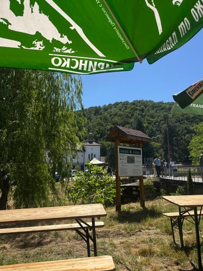 The image shows an outdoor scene in Ustrem, Bulgaria. In the background, a white building with a brown dome and a red roof is visible, likely a religious structure. Behind the building, a forested hill rises against a clear blue sky.

In the midground, an information sign with a wooden roof and green text is present. Several people are visible on a bridge or walkway behind the sign. The foreground is dominated by the partially visible green umbrellas advertising "SHNICKO" and "KAMENITZA TRAKIA". A wooden picnic table is partially visible in the foreground. The ground is covered in grass and some dry patches. A large willow tree with abundant foliage is present. The overall environment suggests a park or recreational area near the building in Ustrem.