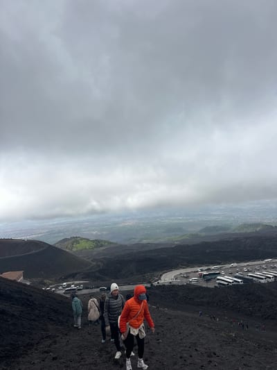 The image depicts a group of people standing on a dark, volcanic slope on Mount Etna, in Nicolosi, Italy. A road with parked buses and cars runs parallel to the slope. The sky is overcast, and a haze obscures the view of the landscape below. There are four people visible in the foreground: one wearing an orange jacket, one in a gray jacket and white hat, one in a beige jacket, and another in a light blue jacket. They appear to be tourists. A building is partially visible at the bottom edge of the image.