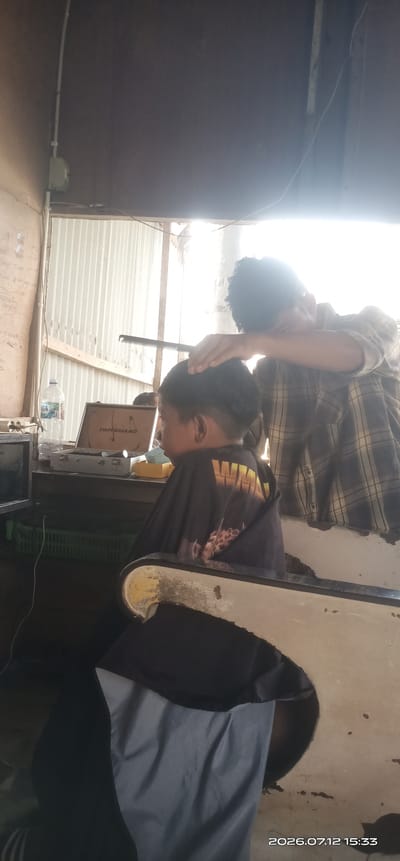 The image depicts a haircut being given to a young person in what appears to be an informal barbershop. The client, seated in a chair, is wearing a black cape with a graphic print. The barber, standing behind the client, is holding a comb and appears to be in the process of cutting the client's hair. The background reveals a somewhat cluttered environment with a corrugated metal wall, a desk or counter with various items including a water bottle, a toolbox, and what looks like a lamp. A white basket is also visible. The lighting suggests it might be daytime, with some bright light entering from the right. The image is tagged with a date and time: 2026.07.12 15:33. The location is identified as Madat, Indonesia.