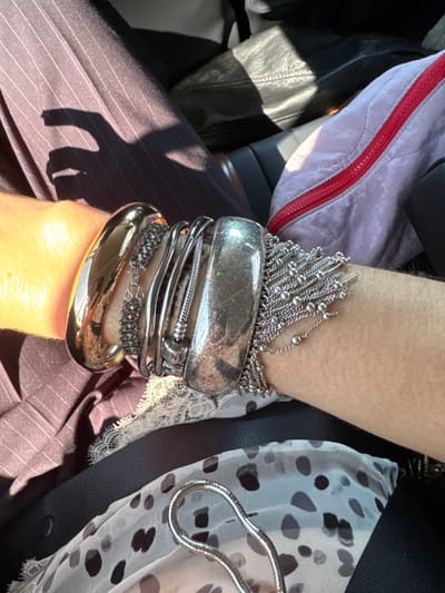 A person's wrist is adorned with multiple bracelets. The top bracelet is silver chains with beads. Below it, a wide, hammered silver bangle. Underneath are several thin, silver bracelets, one a charm bracelet. At the base, a polished gold bangle. The arm rests against pinstripe fabric. To the left are items with pink and red and shiny black surfaces. To the right, a black top with white fabric featuring black dots and white lace edging.