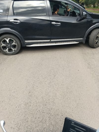 A black compact SUV, likely a Honda BR-V, is observed stationary or moving slowly on a grey asphalt road in Cangkuang Kulon, Indonesia. A female individual with dark hair tied back is seated in the driver's seat, visible through the partially reflective side window, with her hands on the steering wheel. The car features chrome door handles, silver side steps, and multi-spoke alloy wheels, with a Honda logo visible on the left front wheel. The vehicle's exterior shows signs of dust or dirt. In the immediate foreground, a chrome handlebar and a portion of a black rearview mirror from a scooter or motorcycle are visible, indicating the perspective from which the image was captured. Trees and foliage are visible in the background, above and through the car windows.