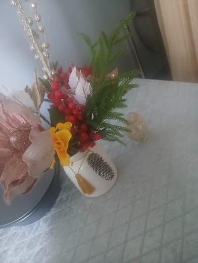 The image shows a decorative arrangement of artificial flowers and berries in a white vase. The vase is decorated with a pine cone design. A yellow flower is prominent in the arrangement, along with red berries and white poinsettia-like flowers. There are also green branches and glittery decorative elements. The vase is placed on a light-colored surface with a patterned texture. The background is blurred and appears to be a wall and possibly a curtain. There is no indication of the city of Khoronk, Armenia. The image is an indoor still life composition.