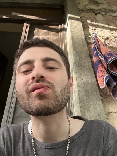 A man with dark hair and a beard is seen from a close-up, upward angle. He wears a gray shirt and a silver chain necklace. Behind him, there is a wall and a folded item, possibly a rug or towel. The scene appears to be indoors near a window or doorway, with light streaming in. The location is Agarakadzor, Armenia.