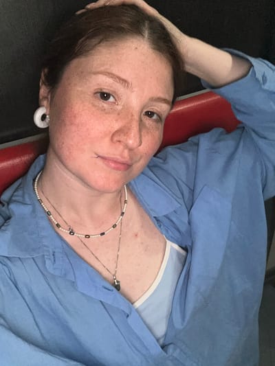 A fair-skinned person with freckles, brown hair, and brown eyes lies on a red surface, possibly a bench or chair, while wearing a light blue button-down shirt partially open to reveal a light blue and white top underneath. The person has a labret piercing below their lower lip and a white hoop earring in their ear. They are wearing two necklaces, one with small beads and another with a thin chain. Their arm is raised behind their head. The environment is dark, and there is no evidence to suggest this image was taken in Vyazniki, Russia.
