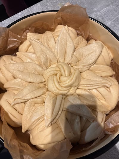 An unbaked, light beige dough is meticulously arranged in a round enamel baking dish, lined with crinkled brown parchment paper. The dough forms a decorative, multi-layered floral pattern, featuring a tightly coiled knot at its center, surrounded by two concentric rings of elongated, petal-like shapes. The surface of the dough appears smooth and slightly glossy, possibly from an egg wash or oil. The baking dish has a cream-colored interior and a distinct black rim. It rests on a grey, textured surface, likely a kitchen countertop. In the upper left background, a blurred dark red or purple object is partially visible. The scene depicts an indoor environment, most likely a kitchen, but provides no visual information to ascertain its specific geographical location within Groß-Gerau, Germany, or any other region.