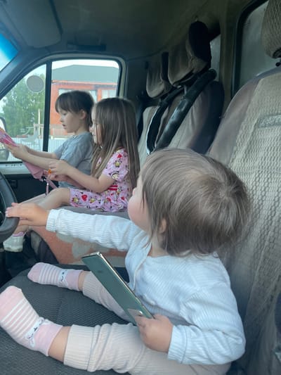 Two young children are in the cab of a vehicle. A younger child is seated on the lap of an older child. The younger child holds a mobile phone. The older child holds a pink object with a handle. Outside the vehicle's window is visible a building and vegetation. The street sign “ул. Строительная, 6а” is seen on the buildings, which translates to “Building Street, 6a”. A sign in Russian is also visible in the windshield. The city is Verkh-Katunskoye, Russia.