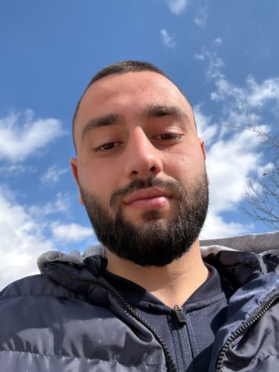 An adult male is depicted in a low-angle outdoor portrait, looking directly forward into the camera. He possesses a short, dark buzz cut and a full, dark beard. His eyes are dark, and his expression is neutral. He is attired in a dark blue puffer jacket layered over a dark blue zippered garment, with a gray fabric lining visible around the neck area, consistent with a hood. The background consists of a clear blue sky interspersed with scattered white cumulus clouds, indicating a bright, sunny day. Bare tree branches are partially visible against the sky in the upper right quadrant. The scene is situated outdoors in Borovichene, Bulgaria.
