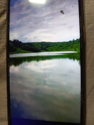 The image shows a landscape scene captured on a cracked phone screen. The scene depicts a body of water with reflections of a cloudy sky and a lush, green forested hillside in the background. The water appears calm, with gentle ripples. The sky is partly cloudy, with variations in light and shadow. The overall impression is of a natural, serene environment. The phone screen itself has several visible cracks, particularly in the upper and central portions of the image, as well as along the right edge. The image is a photograph of a photograph, with the phone screen overlaying the landscape. There is no discernible information about the specific location in Svoboda, Russia, or any people present.