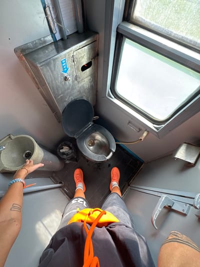 This is a point-of-view photograph taken inside a small, utilitarian train bathroom. The camera is positioned as if the photographer is standing, looking down at their own feet and the immediate surroundings. The space is narrow and appears to be constructed from gray metal and plastic.

Dominating the center of the frame is a black toilet with its lid up. To the left of the toilet is a small, light-colored sink with a faucet. The sink appears to be made of a speckled material. Behind the toilet, a black trash bag is visible.

Above the toilet, a metallic cabinet is mounted on the wall. A blue sticker with Vietnamese text and an arrow pointing down is affixed to it. A clipboard with papers is clipped to the top of this cabinet.

To the right of the toilet, a window offers a view of a bright, overcast sky or landscape, suggesting it is daytime. The window has a gray frame and appears slightly fogged or dirty.

The photographer's legs are visible in the lower half of the image. They are wearing dark gray shorts with an orange drawstring, orange athletic shoes, and white socks. The photographer's left arm is also visible, adorned with a braided bracelet and a tattoo on the forearm consisting of three horizontal lines within a rectangle. Their hand is reaching out towards the sink area, as if interacting with it.

The floor is a dark, tiled surface. A metal handle is mounted on the wall to the right, below the window.

The image captures a very functional and somewhat cramped environment, typical of a train carriage. The inclusion of the person's feet and arm suggests an attempt to document the experience of using this specific facility.