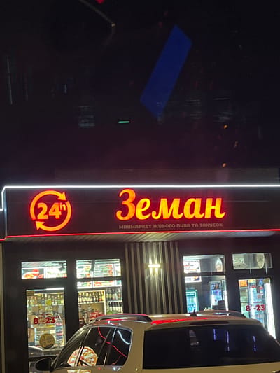 A nighttime exterior shot shows a storefront for a mini-market named "Земан" (Zeman) in Strumivka, Ukraine. The sign is illuminated with bright orange neon letters that spell out the name, and a circular "24h" symbol is visible to the left. Below the main sign, smaller text reads "МІНІМАРКЕТ ЖИВОГО ПИВА ТА ЗАКУСОК" (Mini-market of live beer and snacks). The storefront itself has large windows displaying various products, including bottles of alcohol and what appear to be snacks. A white car is parked in front of the store, and its windshield reflects a pizza. The environment is dark, with the store's lighting being the primary source of illumination.