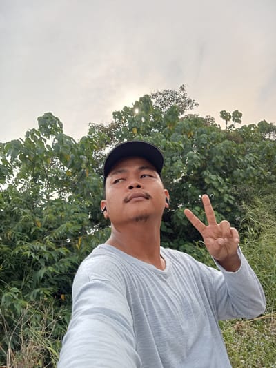 A man wearing a grey long-sleeved shirt and a black baseball cap is standing outdoors in front of lush green trees. He is looking upwards and to the right of the camera, with his right hand raised and making a peace sign. He is wearing black wireless earbuds in both ears. The background consists of dense foliage and grass, with a bright, overcast sky above. The lighting suggests it might be late afternoon or early morning. The man appears to be in a rural or natural setting, possibly outside of Teluk Kenidai, Indonesia.