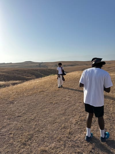 Two people are standing on a barren, dry hillside with a sparsely vegetated landscape. The sky is clear and blue. The person in the foreground is wearing a white t-shirt, black shorts, white socks, and blue running shoes, and has a colorful headdress. The second person, farther away, also wears white clothing. The scene suggests a dry, rural location possibly near Sidi Badhaj, Morocco, but specific landmarks confirming the exact city location are absent.