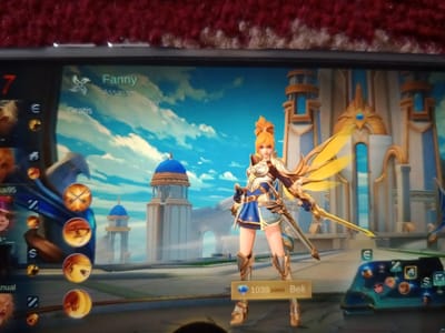 A smartphone screen displaying a mobile video game interface is presented in a close-up photograph, resting on a textured, reddish-brown surface. The central focus is a female game character named "Fanny," identified as an "Assassin" and "Chase/Finisher," also listed as "Gratis" (Free). Fanny has orange hair, fair skin, and wears blue and white armor accented with gold, featuring prominent golden wings and holding a golden sword in a dynamic pose. The game's background depicts a fantasy cityscape under a bright blue sky with white clouds, showcasing white and gold structures, including some with blue domes. On the left side of the screen, a vertical column displays multiple circular icons, likely representing skills or other characters. At the bottom center, a purchase price of "1039 1099" is visible next to a blue diamond icon, followed by the word "Beli" (Buy). The lower right area of the screen shows a control panel or skill bar with various circular icons. This scene is located in the city of Neuheun, Indonesia.