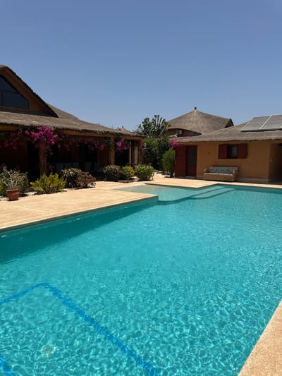 The image shows a building with a thatched roof and solar panels in the Communauté rurale de Malicounda, Senegal. A turquoise swimming pool runs along the side of the building. The building is decorated with flowering plants. The sky is clear and blue.