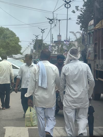 In Datala, India, on an overcast day, four men are seen walking on a street. Two of the men are in the foreground, facing away from the camera. The man on the left is wearing a light-colored, possibly white, long-sleeved shirt and dark pants. He is carrying a translucent yellow plastic bag filled with items. The man to his right is wearing a white checkered long-sleeved shirt and white pants, with a white cloth wrapped around his head. He is carrying a closed black umbrella. In the mid-ground, two other men are visible. One, on the left, is wearing a cream-colored long-sleeved shirt and dark pants. The other man in the mid-ground, partially obscured, is wearing a white shirt. Behind them, a white car is parked on the left side of the street, and a large truck is on the right. The street is paved and has a pedestrian crossing marked by white lines. The background features numerous utility poles with street lights and wires, along with trees and buildings, suggesting an urban or semi-urban environment.