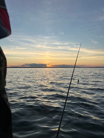 The image depicts a serene sunset scene over a body of water, possibly a lake or sea, with a fishing rod in the foreground. The sun is setting on the horizon, casting a warm, golden glow across the sky and water. The sky is a gradient of blue and orange, with scattered clouds. The water is calm, with gentle ripples reflecting the colors of the sunset. A fishing rod is positioned diagonally in the frame, with a lure hanging from the line. The lower left of the image shows a dark, out-of-focus object, possibly part of a boat or person. The location is identified as Kostovaty, Russia, suggesting a coastal or lakeside setting within this area. There are no visible people actively engaged in any interactions, only the solitary fishing rod suggesting a person is present but not within the frame. The overall atmosphere is peaceful and tranquil.