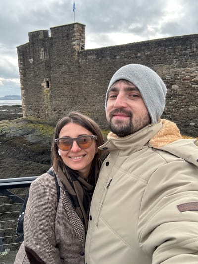 The image depicts a man and a woman in the foreground, posing for a selfie in front of Blackness Castle, located on the southern shore of the Firth of Forth in Blackness, West Lothian, Scotland, United Kingdom.

The man, positioned on the right, wears a light grey knit beanie and a beige insulated jacket with a tan faux fur collar; a "FASHION NORDIC STYLE" patch is visible on his left sleeve. He has a beard and is smiling. The woman, to his left, has long brown hair, wears brown-tinted sunglasses, a white earbud in her right ear, a brown and black patterned scarf, and a brown houndstooth or plaid blazer. She is smiling broadly.

Behind the individuals, the rugged stone walls of Blackness Castle dominate the midground. The castle wall is constructed from dark, irregular stone. A section of a square tower with battlements and narrow embrasures is visible on the left, topped by a flagpole flying a blue flag, likely the Scottish Saltire. Below the castle, the dark, rocky shoreline of the Firth of Forth is visible, with patches of green or dark marine growth. The water beyond the rocks appears calm, and a faint landmass is discernible in the far distance on the left. The sky is overcast with dense grey clouds. A dark metal railing is partially visible in the lower-left foreground, separating the subjects from the immediate shoreline.
