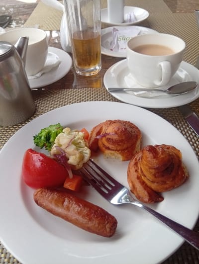 A white plate holds breakfast: a sausage, tomato wedge, steamed vegetables, and two cinnamon rolls. A fork rests on the plate, near the sausage. Behind the plate, a cup of tea sits on a saucer with a spoon. To the left, a silver teapot and another teacup with a saucer are visible. A tall glass of juice and a sugar packet are also present on the table. The breakfast is set on a woven placemat.
