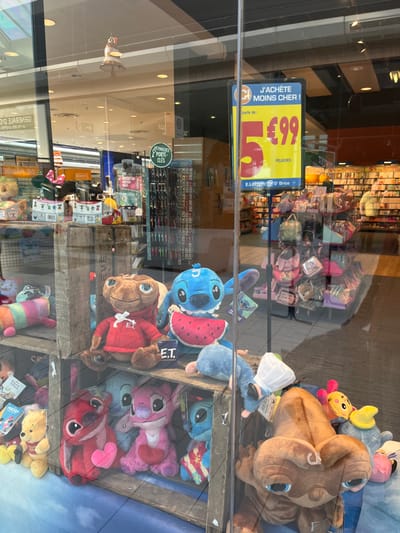 The image shows a storefront display with a variety of plush toys. The storefront is located at 49.283, 4.001 in the city of Saint-Brice-Courcelles, France. The storefront is a store for children's items, such as plush toys, and it is an E.Leclerc store. The display is in the window and is visible from outside. The toys are displayed on wooden shelves, with some plush toys hanging from a rack. The toys are arranged in a random order. Some of the toys are recognizable characters, such as ET and Stitch. The toys are mostly in shades of blue, pink, and brown. The storefront window has a sign advertising plush toys for sale. The sign says "J'achete moins cher!" with a price tag of "5.99."