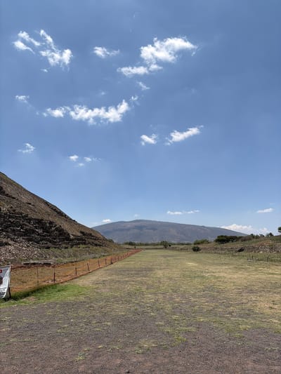 The image depicts a sunny, mountainous landscape in San Sebastián Xolalpa, Mexico. A partially visible ancient Mesoamerican pyramid of Teotihuacan occupies the lower left of the frame. A fence runs alongside the base of the pyramid and extends into a dry, grassy field with rocky terrain. Above the pyramid and field is a clear blue sky with scattered white clouds. A mountain range forms the backdrop to the field.
