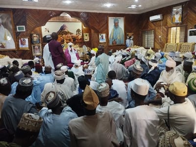 The image depicts a gathering of people, likely in a mosque or religious building, in Kaltungo, Nigeria. They are seated on the floor in rows, facing a central figure seated on a platform. The central figure is wearing a white robe and a white cap. The surrounding people are mostly men, wearing similar white clothing and various forms of white headwear. The environment features wooden walls and ceiling, with framed portraits on the walls. The room appears to be dimly lit, with some overhead lighting. The image captures a moment of religious gathering or a community event, with a focus on the central figure who appears to be leading the group.