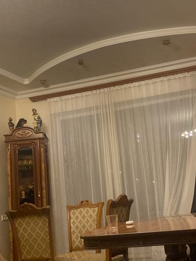 The image depicts an indoor scene, likely a dining room or living area, in Ptghni, Armenia. The room features a ornate wooden cabinet filled with glassware, topped with decorative figurines of a dog and two human-like figures. In front of a sheer, floor-to-ceiling curtain, a polished wooden table is set with a glass and what appears to be a small electronic device, possibly earbuds. Two upholstered chairs with intricate patterns are visible, one partially obscured by the table. The ceiling has a decorative cove with recessed lighting fixtures. The overall impression is one of a well-furnished, traditional interior space.