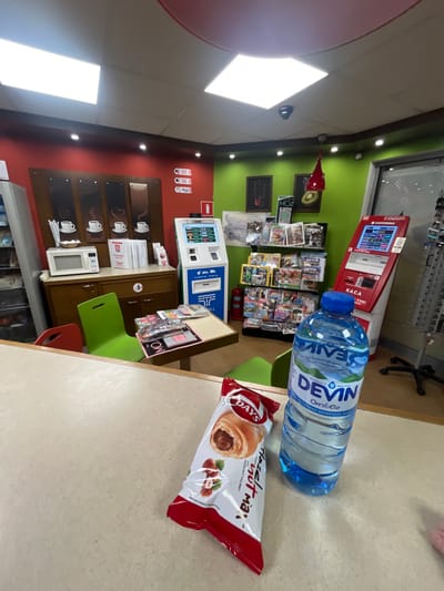 The image depicts the interior of a small shop or convenience store in Momin Sbor, Bulgaria. In the foreground, on a light beige speckled countertop, rests a clear plastic bottle of "DEVIN" mineral water with a blue cap, positioned upright. Next to it is a red and white plastic package of "DAYS Hazelnut max," featuring an image of a croissant with hazelnut filling.

The background shows a brightly lit retail space with distinct wall colors: a deep red on the left half and a vibrant lime green on the right. The ceiling is white with multiple square LED panel lights and recessed spotlights. A red circular ceiling feature is visible. Surveillance cameras are mounted on the ceiling.

Against the red wall, a brown wooden cabinet houses a white microwave oven. Above the cabinet, four dark brown panels with white coffee cup illustrations are mounted. To the right of the cabinet, a blue and white self-service terminal, labeled "A1 Top Up," stands on the floor. Further right, against the green wall, a black metal display rack is stocked with various magazines and newspapers. Adjacent to the rack, a red self-service terminal labeled "KACA" is visible.

In the midground, a small square table with a light brown top is surrounded by two bright green chairs and one red chair. Various items, possibly magazines or small products, are arranged on this table. A red fire extinguisher is visible on the floor near the blue self-service terminal. Decorative elements on the green wall include framed pictures and a hanging red light fixture. No people are visible in the scene.