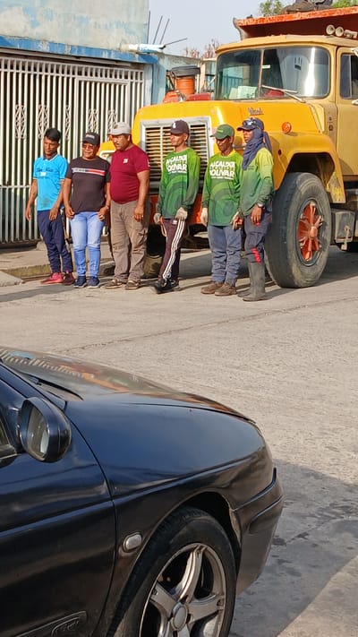 The image displays a group of approximately eight to nine men standing outdoors on what appears to be a paved street or roadside, under bright, clear daylight. The scene suggests a sunny day with distinct shadows.

In the foreground and center, several men are gathered. Three individuals are prominently featured wearing matching green long-sleeved shirts with the text "MCALDIA BOLIVAR" printed in white, dark pants, and baseball caps. One of these men also wears a dark head covering under his cap. Behind them, another man wears a red t-shirt and light-colored pants. Further to the left, two more men are visible, one in a black t-shirt and light blue jeans, and another in a light blue t-shirt and dark pants. The men appear calm, with generally neutral expressions, possibly posing for a photograph or during a work break.

To the left, a large, vintage yellow heavy-duty truck dominates the background. The word "Mack" is visible on its grille. Part of its large front wheel is also seen. The truck's presence, coupled with the "MCALDIA BOLIVAR" uniform, suggests a connection to municipal services or public works in Barcelona, Venezuela, as "Alcaldía de Bolívar" refers to a municipal administration.

On the far right, the front and side of a black car are partially visible, including its wheel with a silver rim and a headlight. In the left background, there's a light blue building wall and a white metal gate with horizontal slats. The overall setting is utilitarian, possibly an industrial or residential area where such work vehicles and teams would operate.