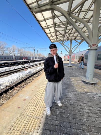 A young man stands on a train platform next to train tracks. He is wearing a black jacket, grey sweatpants, white sneakers, and a black baseball cap. He holds a white object. The platform is made of paving stones. Overhead, there is a metal structure. In the background, a train is visible, along with another person standing near a pillar marked with the number 52. The sky is clear and blue. Bare trees are visible beyond the train. The scene appears to be outdoors on a sunny day. The location is Tashevka, Russia.