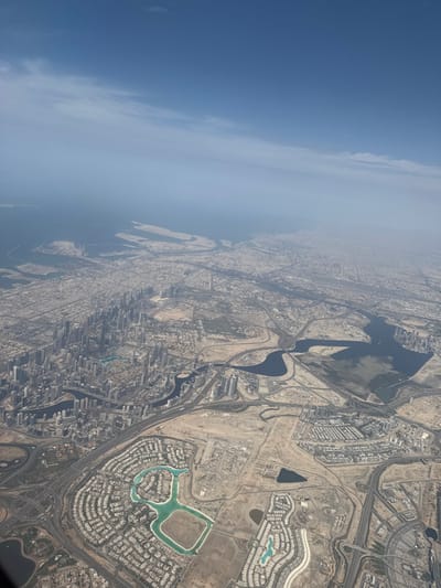 An aerial photograph captures a vast urbanized coastal landscape under a hazy blue sky with faint, wispy clouds. The Persian Gulf occupies the upper left and center, its shoreline extensively modified by numerous artificial islands and land reclamation projects.
Below, the cityscape of Dubai, United Arab Emirates, unfolds. A dense concentration of high-rise buildings forms a prominent central business district in the mid-left, bordering a winding waterway identified as the Dubai Creek/Business Bay Canal. This urban core transitions into widespread residential areas characterized by organized street grids and numerous low-rise structures. In the lower-left quadrant, a distinctive residential development features a large, cyan-colored, figure-eight-shaped artificial lagoon surrounded by concentric housing blocks. Additional residential clusters, some with smaller artificial water bodies, are visible in the foreground.
Complex networks of multi-lane highways, interchanges, and arterial roads traverse the entire scene, connecting disparate urban sectors. Large patches of light brown, arid land, indicative of desert terrain, are interspersed among developed areas, some showing signs of earthworks and ongoing construction. No individual people, vehicles, or specific actions are discernible from this altitude.
The coastal developments and land reclamation projects visible in the upper-right section of the image correspond to areas within the city of Deira, specifically the Dubai Islands (formerly Deira Islands), situated on the northeastern side of the metropolis.
