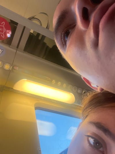 The image shows the interior of a train compartment, likely in the Saran train station in France, located at the coordinates 47.961, 1.891. Two individuals are visible, a man and a woman. The man is in the foreground, his face is close to the camera, showing only his upper body, eyes, nose, and mouth. His head is tilted towards the camera, revealing the side of his face. The woman's head is visible in the lower right corner of the frame, showing her hair, eye, and part of her face. The lighting is fluorescent and casts a yellow hue over the interior of the train compartment. There is a window behind the man and woman, revealing a clear, blue sky with a white cloud. The upper part of the image shows an overhead light, a series of round ventilation openings, and a metal shelf. The man and woman are looking away from the camera, and there appears to be no interaction between them.