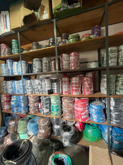 The image captures an indoor storage area or warehouse densely packed with numerous spools of electrical wires or cables, neatly organized on multi-tiered shelves. The shelving unit is constructed from gray metal beams supporting brown wooden planks, creating multiple compartments filled with inventory.
The primary subjects are the coiled wires, which display a vibrant spectrum of colors including red, blue, green, black, brown, purple, light green, and various shades of grey. Most spools are individually wrapped in clear plastic film, indicating new products. Many of these plastic wraps feature white labels with partial text such as "Neutrales," "20 AWG," and "100m," suggesting specifications and lengths. Brand indicators like "DHU" and "AZ" are also visible on some packaging.
Interspersed among the shelves, particularly on the left side, are several cardboard boxes, some open and some closed. One prominent box in the lower left displays the text "TABLEPLAST" along with "VCT328P 150 DCT09." Another box shows "CAJA CPR-070892 PVC."
The setting appears to be under artificial indoor lighting. The photograph is taken from a slightly upward perspective, offering a comprehensive view of the stacked shelves and their contents. The organized arrangement of a large quantity of electrical wires suggests a commercial environment, likely a hardware store or an electrical supply wholesale in Campana, Argentina. No individuals are visible in the scene.