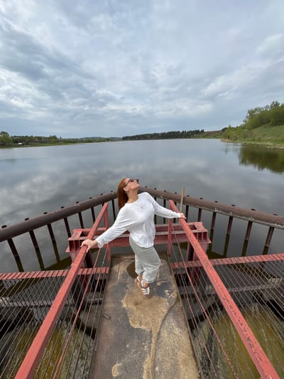 A woman wearing a white long-sleeved shirt and light-colored pants stands on a rusty red metal structure, possibly a bridge or dam, with a body of water visible below and in the background. She is leaning back, holding onto the railing with both hands, and looking upwards. The water reflects the cloudy sky. The opposite bank is lined with trees and some vegetation. The image suggests a rural or natural setting, possibly taken in or near the city of Nozhovka, Russia, given the prompt's specifications.