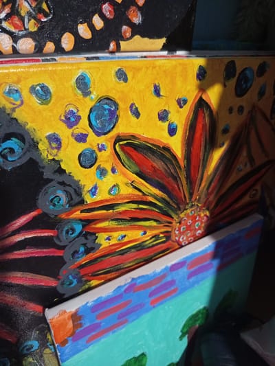 The image displays a close-up, angled view of multiple stacked paintings. The topmost painting, partially visible, is black with colorful, circular splotches of orange, yellow, and white. Below this is a painting with a bright yellow background, decorated with various blue and purple circular shapes, some with darker centers. A prominent feature of this yellow painting is a large, stylized flower with vibrant red, orange, yellow, and black petals radiating outwards. The center of the flower is a textured area of red and green dots. Another painting is visible in the foreground, lying horizontally. This painting features a light blue background with horizontal stripes of pink and purple, and a section of teal with green, abstract shapes. The paintings appear to be acrylic on canvas. The lighting suggests an indoor environment, possibly a studio or storage area, with shadows cast upon the lower painting. The specific location of Чэрвеньскі сельскі Савет, Belarus, cannot be determined from the visual information provided in the image.