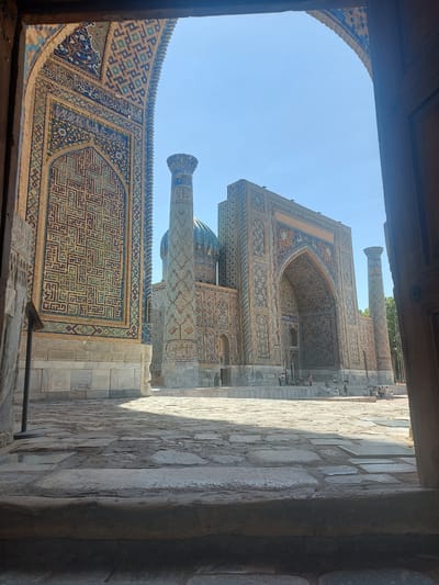 This image shows a view of the Registan complex in Samarkand, Uzbekistan, framed by a large archway. The foreground is dominated by a stone courtyard with large, irregular flagstones. The architecture of the Registan is richly decorated with intricate geometric patterns and calligraphy in shades of blue, yellow, and brown tilework. Two tall minarets stand prominently in front of a large, arched building, which has a turquoise dome visible. Several people are scattered throughout the courtyard and on the steps of the building, appearing to be tourists or visitors. The sky is clear and blue. The scene suggests a sunny day.