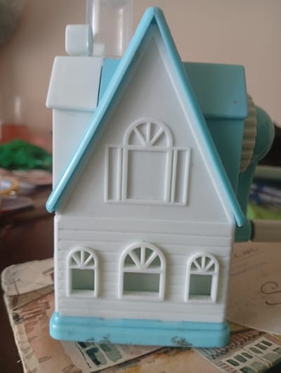 The image shows a close-up, head-on view of a white and light blue plastic pencil sharpener shaped like a house. The house has a light blue, peaked roof with white trim. The front of the house features a larger, arched window with white spokes on the upper level, and three smaller arched windows with similar spokes on the lower level. The lower part of the house is a light blue base. To the right of the house, a portion of the pencil sharpener mechanism is visible, with a ribbed, light brown or beige wheel and a teal colored casing. The object is situated on a surface covered with a patterned paper, possibly old postcards or decorative paper, which has a watercolor effect. The background is blurred and out of focus, with hints of light brown walls and some green and other colors suggesting other objects or surfaces. There are no people visible in the image, and no indication of the specific location within Seýdi, Turkmenistan.