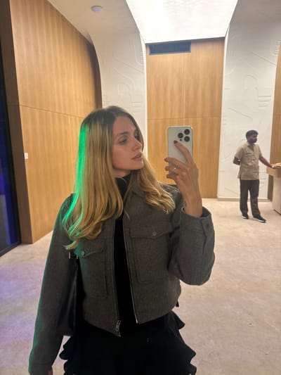A woman with blonde hair is taking a mirror selfie indoors. She is wearing a dark turtleneck, a grey cropped jacket with a zipper and two chest pockets, and a black ruffled skirt. She is holding a silver iPhone in her right hand, and a dark purse is slung over her left shoulder. A man in a tan uniform is visible in the background, standing with his back to the camera. The environment appears to be a modern building with wooden paneling on the walls and abstract white decorative elements on the ceiling and walls. There is a green light cast on the woman's hair from an unseen source. The location is the city of Akhalubani, Georgia.