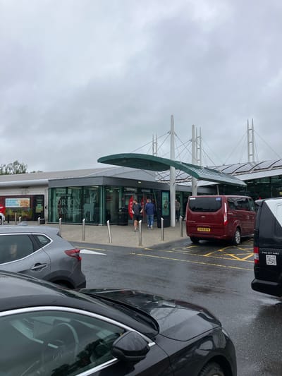 This is a photograph taken from a low angle in a parking lot in front of a modern building with a green awning. The building appears to be a service station or rest stop, with "Welcome to NORTON CANES" visible on a glass facade, indicating the location as Norton Canes, Cannock Chase, United Kingdom. The sky is overcast and the ground is wet, suggesting it has recently rained or is raining.
In the foreground, the hood and windshield of a black car are visible, reflecting the surroundings. To the left of this, a silver car is parked, and a woman is visible in its driver's side window.
In the mid-ground, several vehicles are parked on the wet tarmac marked with yellow lines. A prominent red van with the license plate "SH69 BYN" is parked facing away from the camera. To its right, the rear of a black van with a QR code and the text "FIND OUT MORE" is visible. Two men are walking towards the entrance of the building. The entrance is marked with signs and doors.
The building itself has large glass windows with a silhouette of a deer. The roof of the building is fitted with solar panels. The overall scene is one of a typical overcast day at a roadside service area.