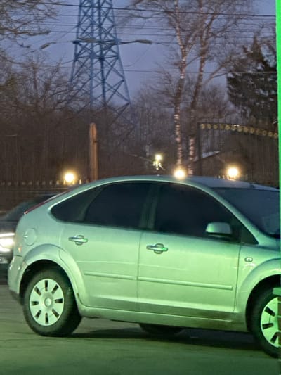 The scene depicts an outdoor setting in 3-Y Uchastok, Russia, under dim, likely evening or night, conditions with a dark blue-grey sky. In the midground, a silver five-door hatchback, consistent with a second-generation Ford Focus, is parked or idling, presenting its left side. Its windows are tinted dark, and the vehicle shows some dirt accumulation on its lower body and wheel wells. Its wheels feature multi-spoke hubcaps. Partial visibility of another vehicle, possibly a darker sedan, is seen to the left and rear of the silver hatchback, with only its front left headlight and bumper visible.
The background features a prominent, light blue or grey metal lattice power transmission tower. Densely packed, bare-branched deciduous trees, including some with distinct white bark, extend across the frame behind the vehicles and the tower, suggesting a cooler season. Several bright, circular yellow-orange light sources are visible within the tree line and above the cars, indicative of streetlights or outdoor lamps. A tall, slender brown vertical pole is positioned behind the silver car. To the far right, an ornate dark metal fence or gate with decorative finials is partially visible, behind which a building structure can be faintly discerned. No people are visible, and no discernible actions or interactions are occurring. The environment presents a mix of natural elements and urban infrastructure.