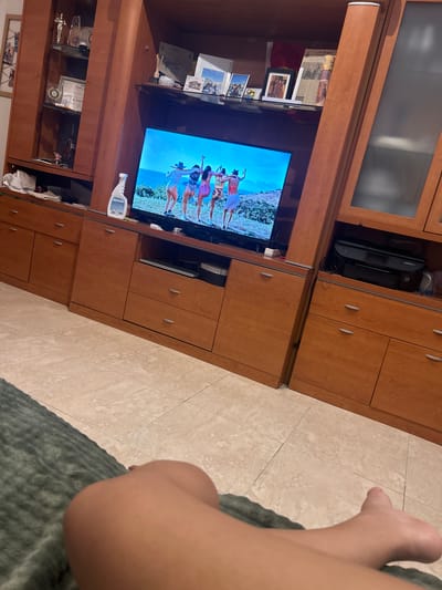 The image shows a person's leg and foot in the foreground, resting on a dark grey, textured blanket. The person is positioned on the right side of the frame, partially obscuring the view of a television screen which is the main focus of the background.

The television displays a group of people in colorful attire, appearing to be dancing or posing on what looks like a beach or coastal landscape. The sky is a bright blue, suggesting it is daytime.

Behind the television is a large, wooden entertainment unit with multiple cabinets and shelves. Some shelves are filled with various objects, including framed pictures, certificates, trophies, and what appears to be a printer. The overall setting appears to be a living room or den.

The lighting suggests it is daytime, possibly late afternoon given the shadows. The presence of a television and home furnishings indicates an indoor domestic environment. No specific emotions are discernible from the image.

There is some visible text in the image, notably on a certificate displayed on a shelf, which includes "TEAM LE" and "$ 2400". A white spray bottle is also visible on the edge of the television stand.

The image is taken from a slightly elevated angle, looking down towards the television and the floor. The floor is tiled with light-colored square tiles. The orientation of the image is tilted to the right. The overall impression is a casual, domestic scene.