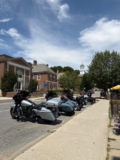 The image depicts a street scene in Peterborough, United States of America, on a bright, cloudy day. In the foreground, several motorcycles are parked along the side of the road. The closest motorcycle is a silver Harley-Davidson, featuring saddlebags and a large front fairing. Behind it, other motorcycles of varying colors are visible, including a dark grey one and several black ones.

The street runs alongside a sidewalk, which has a black metal fence bordering a garden area with purple flowers. A yellow umbrella is set up over a table at what appears to be an outdoor seating area for a cafe or restaurant. A chalkboard sign stands near the entrance to this area.

Across the street, there are several brick buildings. The building on the left has a grand entrance with an arched window and classical architectural details. To its right, another brick building with a gabled roof is visible. Further in the background, a white building with a green steeple and clock tower stands against the sky, which is filled with scattered white clouds against a blue backdrop. Trees are interspersed throughout the scene, with one large tree prominently featured on the right side of the image. An American flag is flying from a flagpole in front of one of the buildings. A few people are casually walking on the sidewalk in the background.