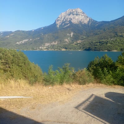 The image shows a wide view of a large, calm, blue lake with a mountain range in the background. The foreground is dominated by dry, yellowish grass and a dirt path, with some green bushes and trees to the left and right. The mountain range is covered in dense green forest, with a prominent, rocky peak at the center. The sky is a clear, bright blue. A small red boat is visible in the distance on the lake, leaving a white wake. In the far distance, along the shoreline of the lake, there appears to be a small town or village with white buildings. The scene suggests a sunny day in a mountainous, natural environment.