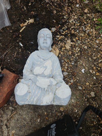 An outdoor scene depicts a light grey Buddha statue resting on an uneven surface composed of dark soil, small stones, and dried organic debris. The statue, rendered in a meditative posture with crossed legs and hands clasped, exhibits areas where an underlying darker grey material is exposed through chipped or worn lighter grey paint. To the lower left of the statue, a single red brick lies horizontally. To the lower right, a black object, possibly a pump or outdoor equipment, is partially visible, displaying white text that reads "Reading Borough Council," indicating the location is within the jurisdiction of Reading, United Kingdom. The ground around the statue is a mix of disturbed earth and gravel, suggesting an uncultivated area. No individuals or active elements are present in the frame.