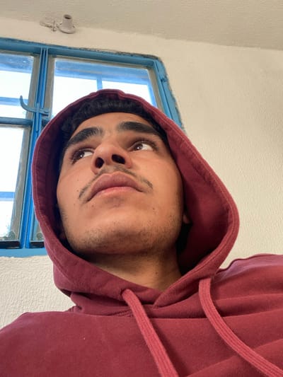 An upward-angled close-up depicts a young male within an interior setting in Dahiyat Harasta, Syria. The subject, positioned centrally, is observed from the chest up, wearing a maroon hooded sweatshirt with the hood drawn over his head. He possesses dark hair, thick eyebrows, dark eyes, and a light mustache with stubble on his chin and cheeks. His gaze is directed upwards and to the right of the frame, with his mouth slightly parted. To the upper left and behind the subject, a window with distressed blue-painted wooden frames is visible, admitting ambient light. Above the window, on the textured white ceiling, a small, white, exposed electrical light fixture base is present, affixed near a patch of peeling paint. The wall to the subject's right is also white and textured.