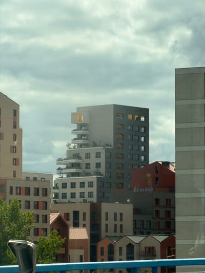 This is a vertical, low-angle photograph of a cluster of modern apartment buildings against a cloudy sky. The buildings are of varying heights and architectural styles, with many featuring balconies and dark-framed windows. Some buildings have red-tiled roofs and brown or grey facades, while others are lighter in color. Green trees are visible in the lower right portion of the image, suggesting a residential or urban park setting. The sky is overcast with soft, diffused light, indicating it could be daytime, possibly late afternoon or early morning, or simply a cloudy day. There is a bright blue pole on the right side of the image, and part of a black security camera is visible in the bottom right corner. The image appears to be a reflection on a window or glass surface, as indicated by a faint reflection of what looks like a ceiling or internal structure at the top of the image. The scene is quiet and appears to depict an urban landscape in Alfortville, France. There is no visible text in the image.