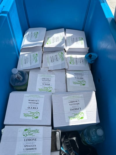 The image shows a close-up, high-angle view of the inside of a blue plastic container, likely a cooler or storage bin. The container is filled with numerous cardboard boxes, most of which are white with green and black text and graphics. The boxes appear to contain individually packaged items, possibly food or beverages, with labels indicating flavors such as "Fragola" (Strawberry), "Anguria" (Watermelon), "Limone" (Lemon), "Menta" (Mint), "Cocco" (Coconut), and "Pesca" (Peach). Some boxes also have text like "Granita Fun" and "SPAGGIÀ".

In the lower left corner, a large plastic bottle of water with a blue cap is visible. To the right of the water bottle and slightly behind it, a dark-colored bag or backpack is partially visible. In the upper right quadrant, a small blue plastic object with a smiley face design is resting on the edge of the container. The lighting suggests an outdoor, daytime setting, likely in a sunny environment, as indicated by the bright illumination and shadows. The blue container itself appears to be in good condition, with some scuff marks visible on its inner surface. The overall scene depicts a collection of packaged goods stored within a portable container, suggesting preparation for an event or transport. The location is identified as Crabonaxa/Villasimius, Italy, which provides a geographical context to the scene.